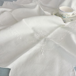 May include: A white, scalloped-edge tablecloth with embroidered floral details. A teacup and saucer with blue floral designs rest on the cloth. The tablecloth is folded, showing the texture and design.