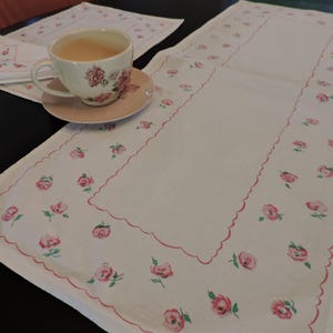 May include: A white table runner, teacup, saucer, and napkin set. The runner and napkin feature a scalloped edge and a repeating pattern of pink roses with green leaves. The teacup and saucer have a matching rose design.