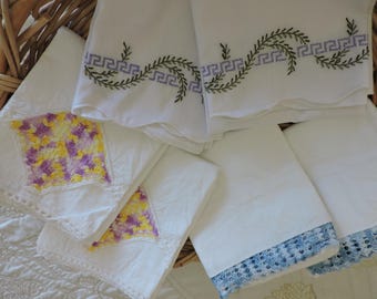Puede incluir: Una colección de ropa de cama blanca con varios acentos decorativos. Algunas piezas muestran bordados griegos en verde y morado, mientras que otras están terminadas con bordes de ganchillo floral amarillo y morado o azul.
