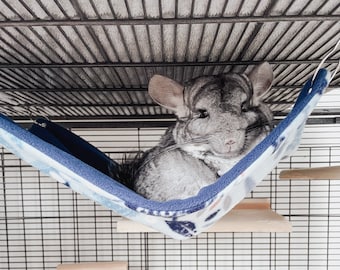 May include: A gray chinchilla rests in a blue and white patterned hammock inside a wire cage. The cage also features wooden shelves at different levels. The chinchilla is the focal point, with its large ears and soft fur.