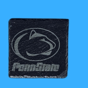 May include: A square, black slate coaster with the Penn State Nittany Lions logo and the text "Penn State" etched in a lighter color. The coaster is set against a bright blue background.