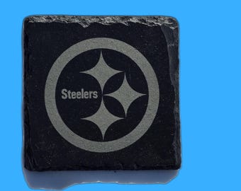May include: Square black slate coaster with the Pittsburgh Steelers logo etched in gray. The logo features a circular design with four star-shaped elements and the word "Steelers" in a sans-serif font.