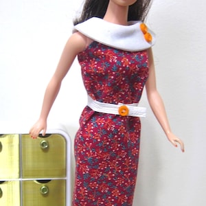 May include: A vintage doll wearing a red floral print dress with a white collar and belt. The dress has a fitted silhouette and is accessorized with an orange button. The doll has blue eyes and red lipstick.