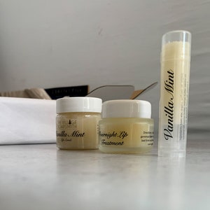 May include: A set of lip care products, including a lip scrub, overnight lip treatment, and a lip balm. The products are in small glass jars and a tube, with labels that read "Vanilla Mint". The background is a neutral white.