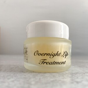 May include: A small, clear jar with a white lid, containing a yellow-tinted lip treatment. The text "Overnight Lip Treatment" is printed on the front in elegant script. The jar is designed for overnight lip care.