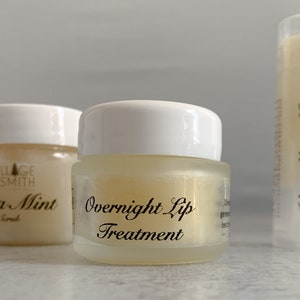 May include: A small, clear glass jar with a white lid, labeled "Overnight Lip Treatment" in elegant black script. The jar contains a pale yellow balm. Another jar and a lip balm tube are visible in the background.