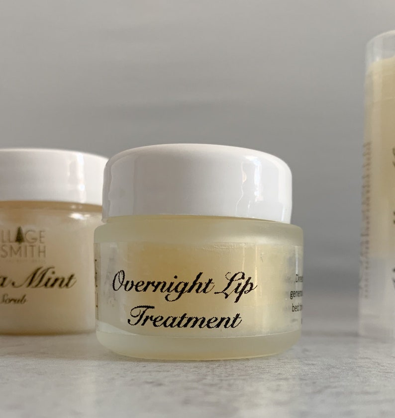 May include: A small, clear glass jar with a white lid, labeled "Overnight Lip Treatment" in elegant black script. The jar contains a pale yellow balm. Another jar and a lip balm tube are visible in the background.