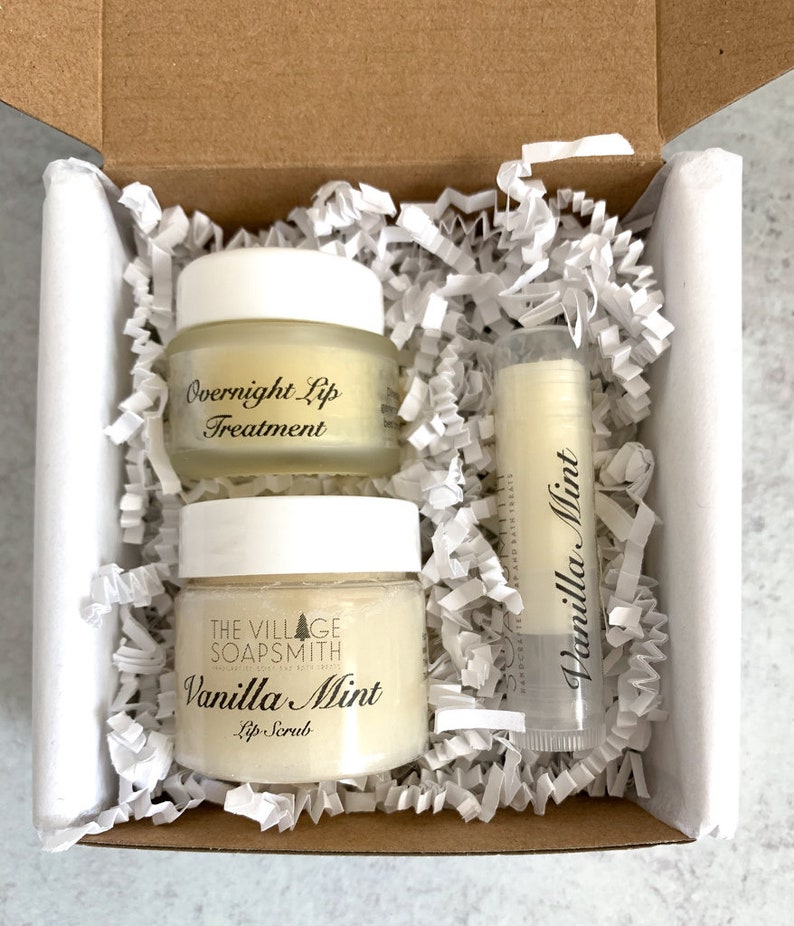 May include: A gift box containing lip care products. The box includes two small jars labeled "Overnight Lip Treatment" and "Vanilla Mint Lip Scrub", and a lip balm stick with "Vanilla Mint" printed on it. The products are surrounded by white paper filler.