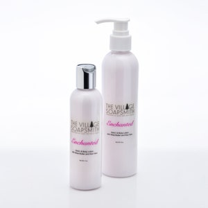 K&ouml;nnte beinhalten: Zwei wei&szlig;e Flaschen Hand- und K&ouml;rperlotion mit Pumpspender. Die Flaschen sind mit "The Village Soapsmith" und "Enchanted" mit Sheabutter und Aloe Vera beschriftet.