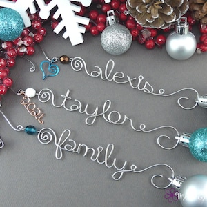 May include: Three silver wire ornaments with the names "Alexia", "Taylore", and "Family" written on them. Each ornament has a small bead and a silver ball at the end. The ornaments are surrounded by red berries and a white snowflake.