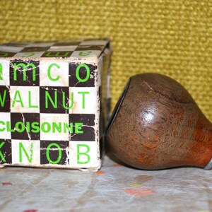 May include: A teardrop-shaped walnut wood knob with a metallic base, positioned next to its original square cardboard box. The box features a black and white checkerboard pattern with the green text: amco WALNUT CLOISONNE KNOB.