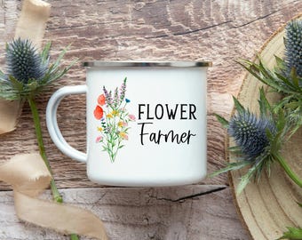 Könnte beinhalten: Ein weißer Emaillebecher mit silbernem Rand, der eine farbenfrohe Aquarellillustration von Wildblumen und den schwarzen Schriftzug "FLOWER Farmer" auf der Seite zeigt. Der Becher hat einen geschwungenen Henkel und eine glatte Oberfläche.