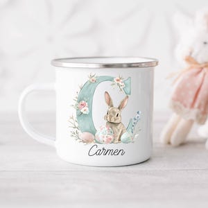 May include: White enamel mug with a silver rim, featuring a watercolour design of a bunny, flowers, and the letter "C" in teal. The name "Carmen" is written below the bunny. A decorative Easter-themed mug.