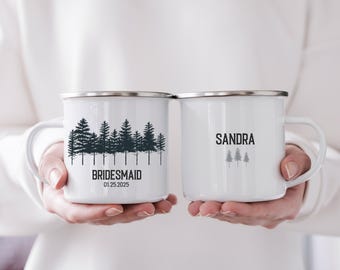 Puede incluir: Dos tazas de esmalte blanco con bordes plateados. Una taza tiene un gráfico de bosque y la palabra "BRIDESMAID" con una fecha. La otra taza tiene el nombre "SANDRA" y un gráfico de bosque más pequeño. Fondo blanco.
