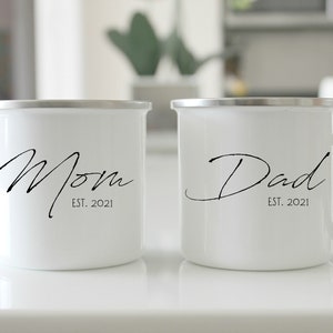 May include: Two white enamel mugs with black lettering. One mug says "Mom Est. 2021" and the other says "Dad Est. 2021".