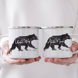 May include: Two white enamel mugs with black bear illustrations and text. The mug on the left says "Uncle Wes est. 2023" and the mug on the right says "Auntie Nina est. 2023".