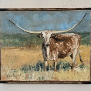 May include: An oil painting of a longhorn steer with a white face and brown body standing in a golden field under a blue sky. The artwork is enclosed in a dark wooden frame and features the artist's signature, Brad Bohl, in the bottom left corner.