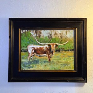 May include: An oil painting of a brown and white Texas Longhorn steer with long, curved horns standing in a green pasture. The artwork is enclosed in a thick, black rectangular frame with gold inner detailing and a signature in the bottom right corner.