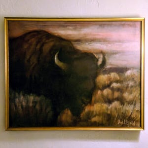May include: Oil painting of a bison in a gold frame. The bison is dark brown with curved horns, set against a backdrop of a pink and tan sky and tan brush. The painting is signed in the lower right corner.