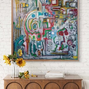 May include: Abstract painting with a colorful, geometric design. The painting is framed in a wooden frame and is hanging on a white brick wall. There are sunflowers in a vase on a wooden table in front of the painting.