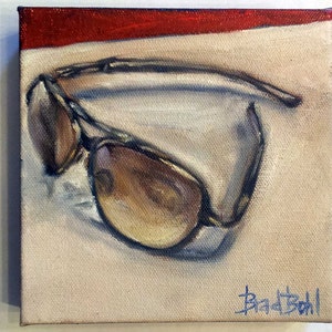 Op de afbeelding: Een schilderij van een paar bruine aviator-zonnebrillen op een beige achtergrond met een rode streep aan de bovenkant. De handtekening van de kunstenaar, "Brad Boll", staat rechtsonder.