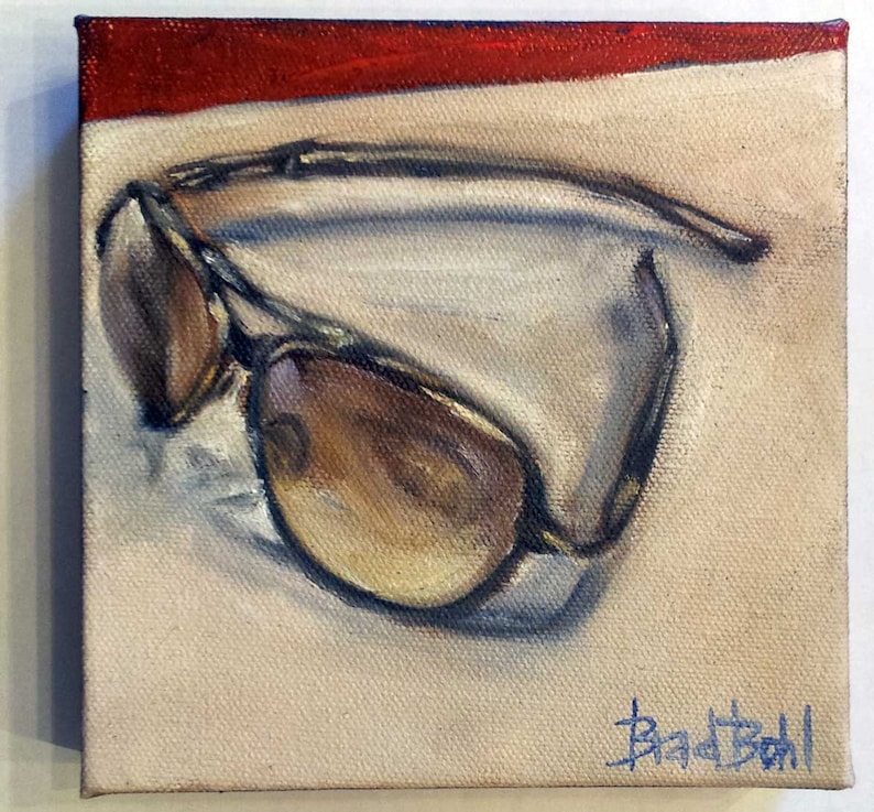 Op de afbeelding: Een schilderij van een paar bruine aviator-zonnebrillen op een beige achtergrond met een rode streep aan de bovenkant. De handtekening van de kunstenaar, "Brad Boll", staat rechtsonder.