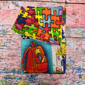 Könnte beinhalten: Ein farbenfrohes Stoffportemonnaie mit einem Puzzleteil-Muster, einem roten Rucksack mit einem Stern und einem Haus mit dem Wort "RU". Das Portemonnaie besteht aus verschiedenen gemusterten Stoffen.