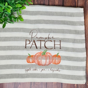 May include: A white and gray striped kitchen towel with the text "Pumpkin Patch" embroidered in brown. The towel also features an embroidered image of three orange pumpkins with green stems and leaves. The text "apple cider. pie. hayrides" is embroidered below the pumpkins.