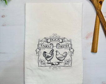 May include: White kitchen towel with black embroidery of two chickens and a banner that reads "EGGS FARM FRESH".