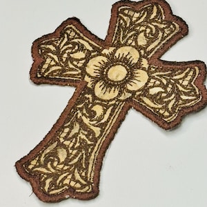 May include: A cross-shaped embroidered patch featuring a tan background with dark brown thread detailing. The design includes intricate swirling vine patterns and a central flower motif, all outlined with a thick dark brown border.