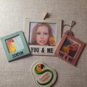 Könnte beinhalten: Eine Auswahl an dekorativen Rahmen und ein Aufnäher mit einem Papageien-Design. Die Rahmen sind in verschiedenen Farben gehalten, darunter Grün, Rosa und Grau, mit dem Text "YOU & ME" auf einigen. Der Aufnäher hat ein Papageien-Design mit dem Text "Suathega Designs".