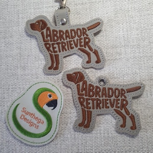 May include: Three embroidered dog-themed keychains and a patch. Two keychains are shaped like Labrador Retrievers with the breed name stitched in brown. The patch features a parrot design with the text "Sunthega Designs".