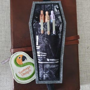 May include: A brown leather journal with a black coffin-shaped pen holder. The pen holder has a black and white cat print and holds three pens. A green and white parrot-shaped patch with the text "Sunthega Designs" is attached.