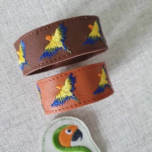 May include: Two leather bracelets with embroidered parrot designs. One bracelet is brown, the other is orange. The parrots are yellow, blue, and orange. A white patch with a parrot design and the text "Synthega Designs" is also visible.