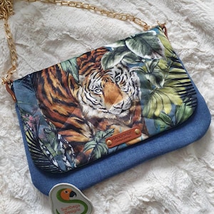 Può includere: Una borsa a tracolla in denim blu con stampa di tigre e foglie tropicali. La borsa ha una tracolla a catena dorata e un'etichetta in pelle marrone che dice "Handmade, limited edition".