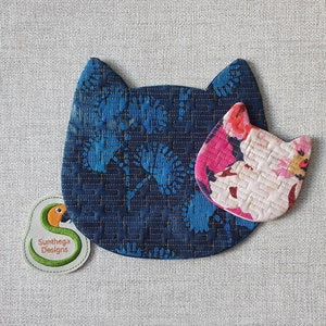 May include: Two quilted fabric cat-shaped coasters. The larger coaster is blue with a floral pattern and gold stitching. The smaller coaster is white with pink and red floral accents. The coasters are on a light gray textured surface. The text "Sunthaga Designs" is embroidered on a green and yellow bird-shaped patch.