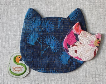 May include: Two quilted fabric cat-shaped coasters. The larger coaster is blue with a floral pattern and gold stitching. The smaller coaster is white with pink and red floral accents. The coasters are on a light gray textured surface. The text "Sunthaga Designs" is embroidered on a green and yellow bird-shaped patch.