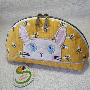 May include: A yellow fabric cosmetic bag with a white bunny face embroidered on the front. The bunny has blue eyes and pink ears. The bag has a zipper closure and a small green and orange fabric tag that says "Sunthaga Designs".