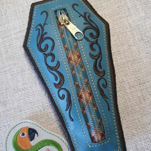 May include: A small blue leather zippered pouch shaped like a coffin with brown embroidered details. The zipper is gold and the pouch is lined with a plaid fabric.