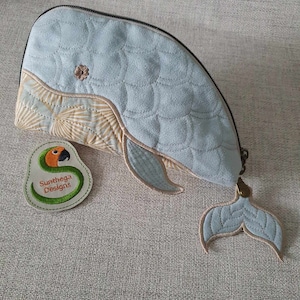 Könnte beinhalten: Eine hellblaue, gesteppte, walförmige Reißverschlusstasche mit einem braunen und orangefarbenen Papageien-Patch, auf dem "Sutthega Designs" steht. Die Tasche hat eine abnehmbare Schwanzflosse.