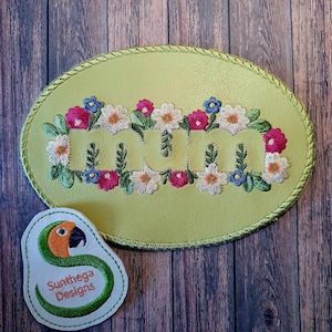 May include: A light green oval-shaped patch with the word "MUM" embroidered in white with a floral border of pink, white, and blue flowers. The patch is surrounded by a green stitched border. A small white patch with a green parrot and the text "Sunthaga Designs" is in the lower left corner.