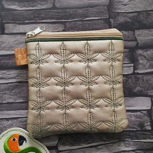 May include: A small, gold-colored zippered pouch with a green trim and a quilted pattern of green leaves. The pouch is sitting on a gray stone background.