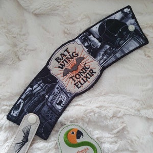 May include: A black and white coffee cozy with a bat wing design and the text "Bat Wing Tonic Elixir". The cozy has a white strap with two white buttons and a black bat design. A green and orange embroidered patch with the text "Sunthaga Designs" is in the lower right corner of the image.