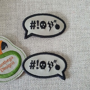 May include: Two embroidered patches in the shape of speech bubbles. The patches are light gray with a dark blue border. Each patch features a skull, lightning bolt, and a bomb graphic, along with the text "#!."