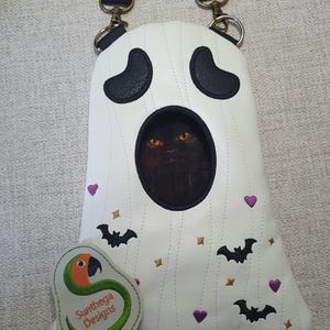 May include: A white ghost-shaped crossbody bag featuring black embroidered eyes and a central circular cutout revealing a black cat with orange eyes. The bag is decorated with small black bats, purple hearts, and gold stars, and includes a black strap.