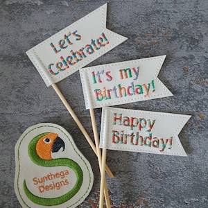 May include: Three white felt flags with colorful embroidery on wooden sticks. The flags read "Let's Celebrate", "It's my Birthday!" and "Happy Birthday!" A white felt patch with a green and orange parrot design and the text "Sunthaga Designs" is in the foreground.