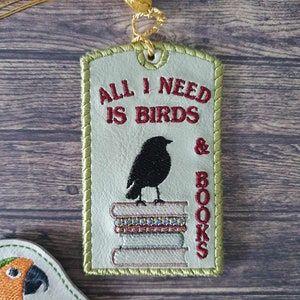 Puede incluir: Un marcapáginas de cuero verde claro con un borla dorada. El marcapáginas tiene un pájaro negro posado sobre una pila de libros y el texto "All I need is birds & books" en bordado rojo.