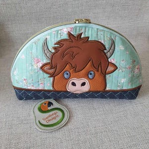 May include: A light blue quilted makeup bag with a brown and white embroidered Highland cow face on the front. The bag has a gold zipper and a green and orange embroidered tag that says "Sunwheya Designs".