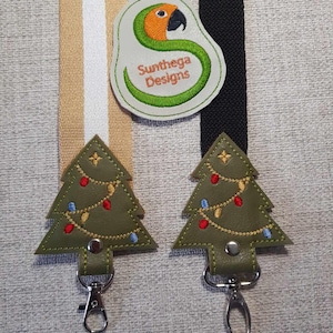 May include: Two green felt Christmas tree keychains with red and blue lights and gold string. Each keychain has a silver metal clasp. The keychains are hanging from a tan and white striped ribbon and a black ribbon. A green felt patch with an orange parrot and the text "Sunthaga Designs" is in the upper center of the image.
