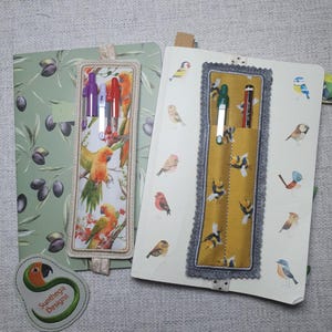 Peut inclure: Deux carnets avec des porte-stylos décoratifs. L'un présente un motif de branche d'olivier, l'autre des illustrations d'oiseaux. Les porte-stylos présentent des motifs d'oiseaux et d'abeilles, contenant des stylos de différentes couleurs. Un écusson de logo est également visible.